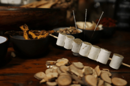 Man sieht mehrere Pflanzenteile in Schüsseln im Hintergrund stehen. Davor sind drei Akupunkturnadeln, die im Tisch stecken und ein Spieß mit darauf aufgereihten Beifußkrautkegeln.